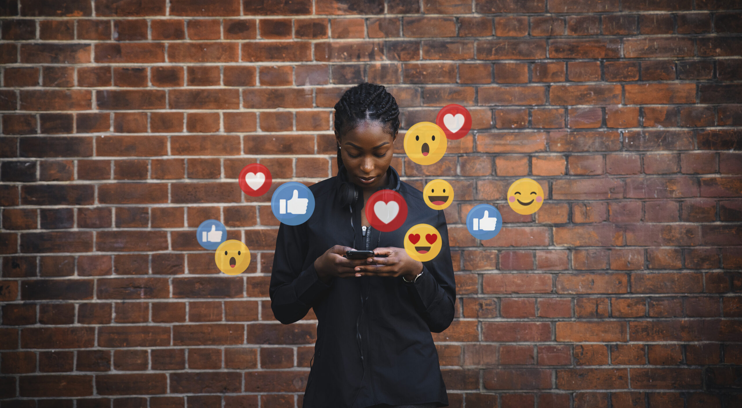 A woman looks at her phone with social media reaction icons floating around her in front of a brick wall.